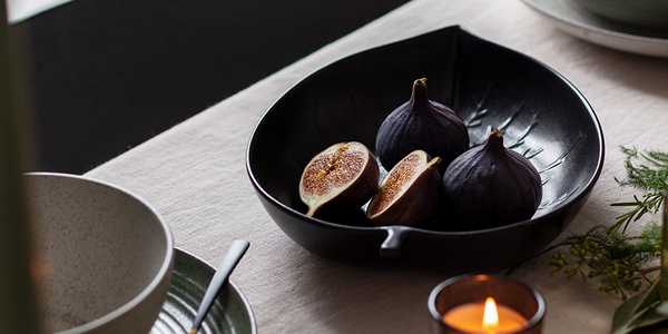 A black leaf shaped serving bowl on a dining table with figs in it. 